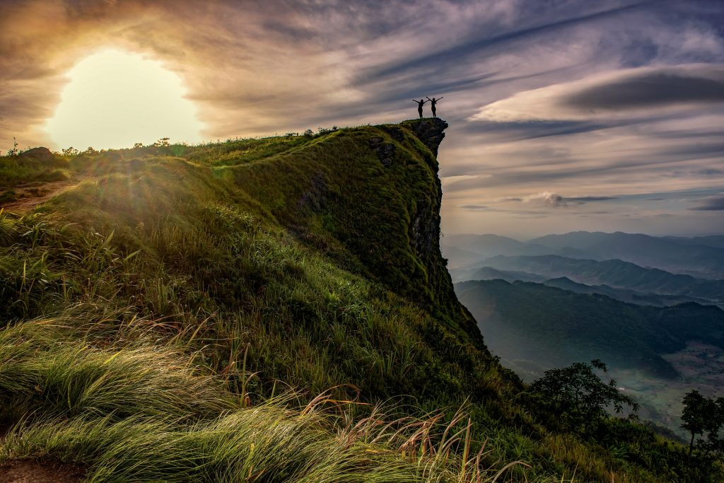 Erfolgreicher Aufstieg - ©sasint/Pixabay Zwei Personen im Sonnenuntergang stehen auf einem hervorstehenden Felsen und strecken jubelnd die Arme nach oben.