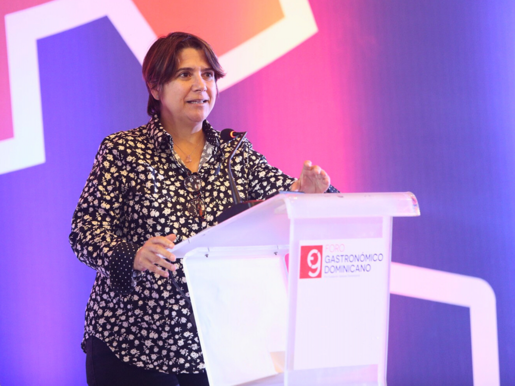 In the picture, Lisette is standing behind a lectern in front of a wall illuminated with orange and purple lights.
